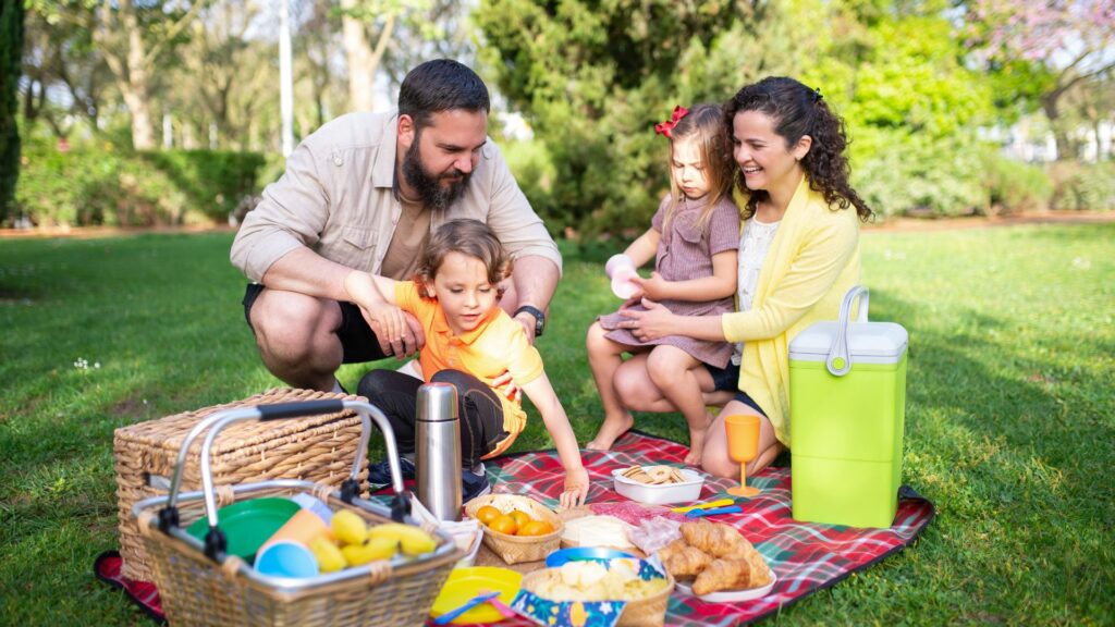 A family of four with a mom and a dad, a son, and a daughter enjoying a picnic in the park together on a red and green plaid blanket with baskets, a cooler, and plates of food on the blanket.