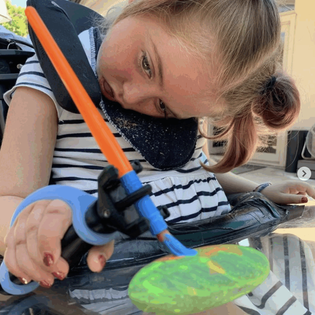 Young girl using the functionalhand® with an Eazyhold® silicone grip to hold a paintbrush so she can paint rocks. Painting is an excellent adaptive summer activity.