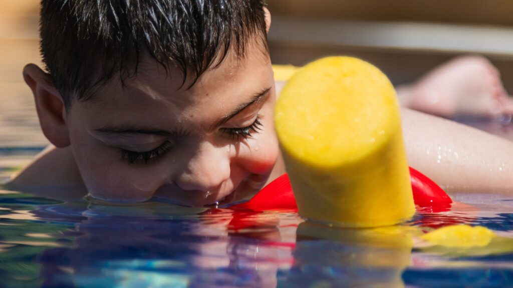 One of our all-time favorite summer activities is swimming. This is a photo of a boy with dark hair holding a yellow pool noodle while blowing bubbles in the water.
