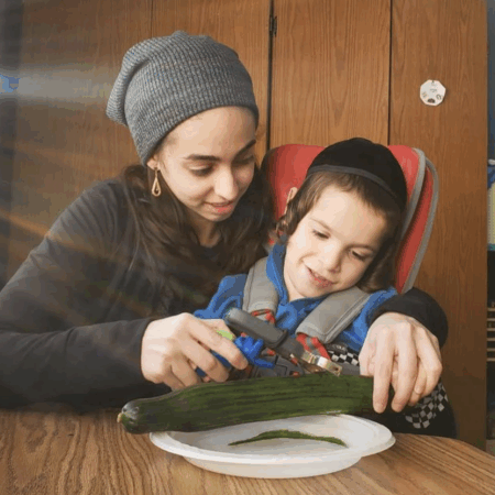 Adult and child using the FunctionalHand assistive device to hold a vegetable peeler and prepare food during daily kitchen routines.