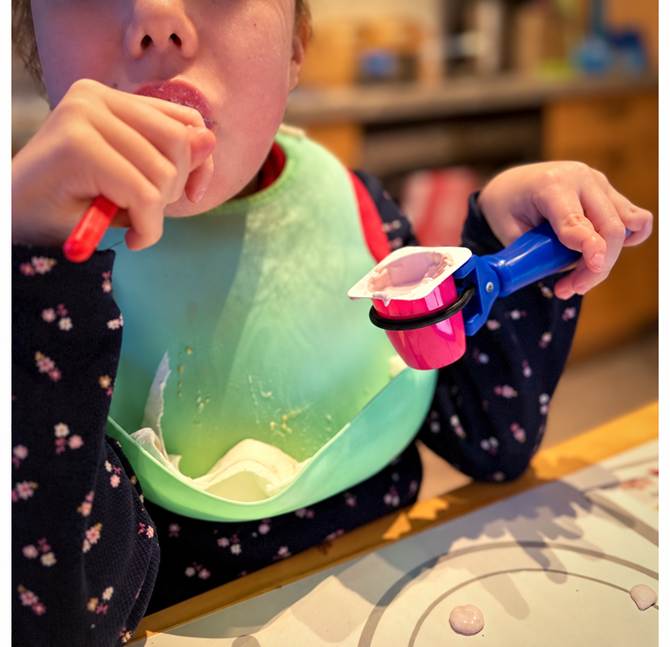This is an image of a girl feeding using a spoon and the functionalhand is holding the food container so the girl can use two hands for the task.