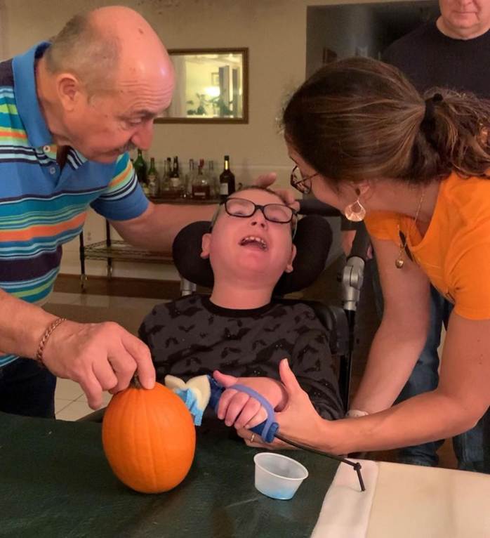 Inclusive Halloween activity for child. Father and therapist helping teen age boy paint pumpkin with functionalhand and eazyhold assisting to hold brush.