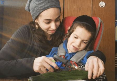 Adult and child using the FunctionalHand assistive device to hold a vegetable peeler and prepare food during daily kitchen routines.